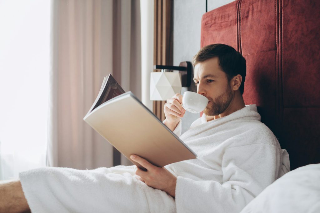 A handsome young man in a bathrobe rests on a bed in a hotel room.